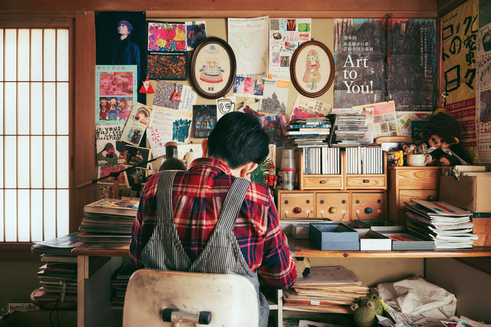 【写真】自宅内のアトリエで、机に向かい絵を描く田湯さんの後ろ姿。机の上やまわりには、ノートやスケッチブックが積み重なり、壁には展覧会のポスターなどがびっしりと貼られている。