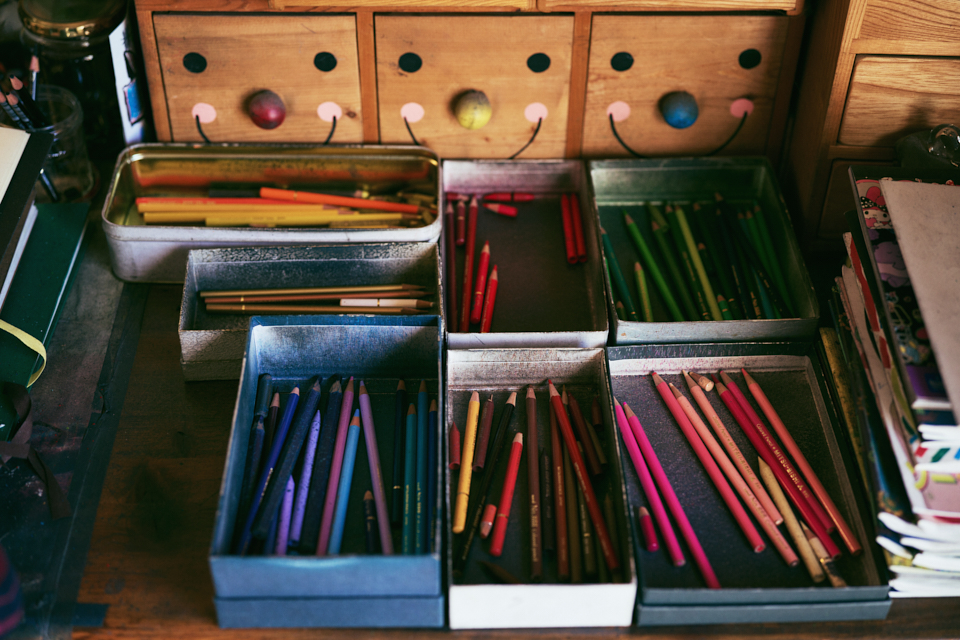 【写真】机上に色鉛筆が入った箱や缶ケースが並んでいる。田湯さんが作品制作に使っているもので、同系色ごとに分けて収納されている。