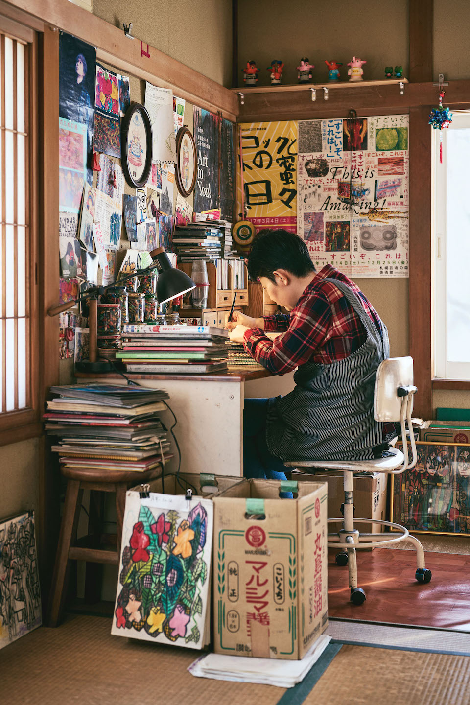 【写真】自室の机で黙々と絵を描く田湯さんの姿。机の脇には絵がクリップで留められたダンボールや、額装された作品、丸椅子の上にうずたかく積まれたスケッチブックがある。