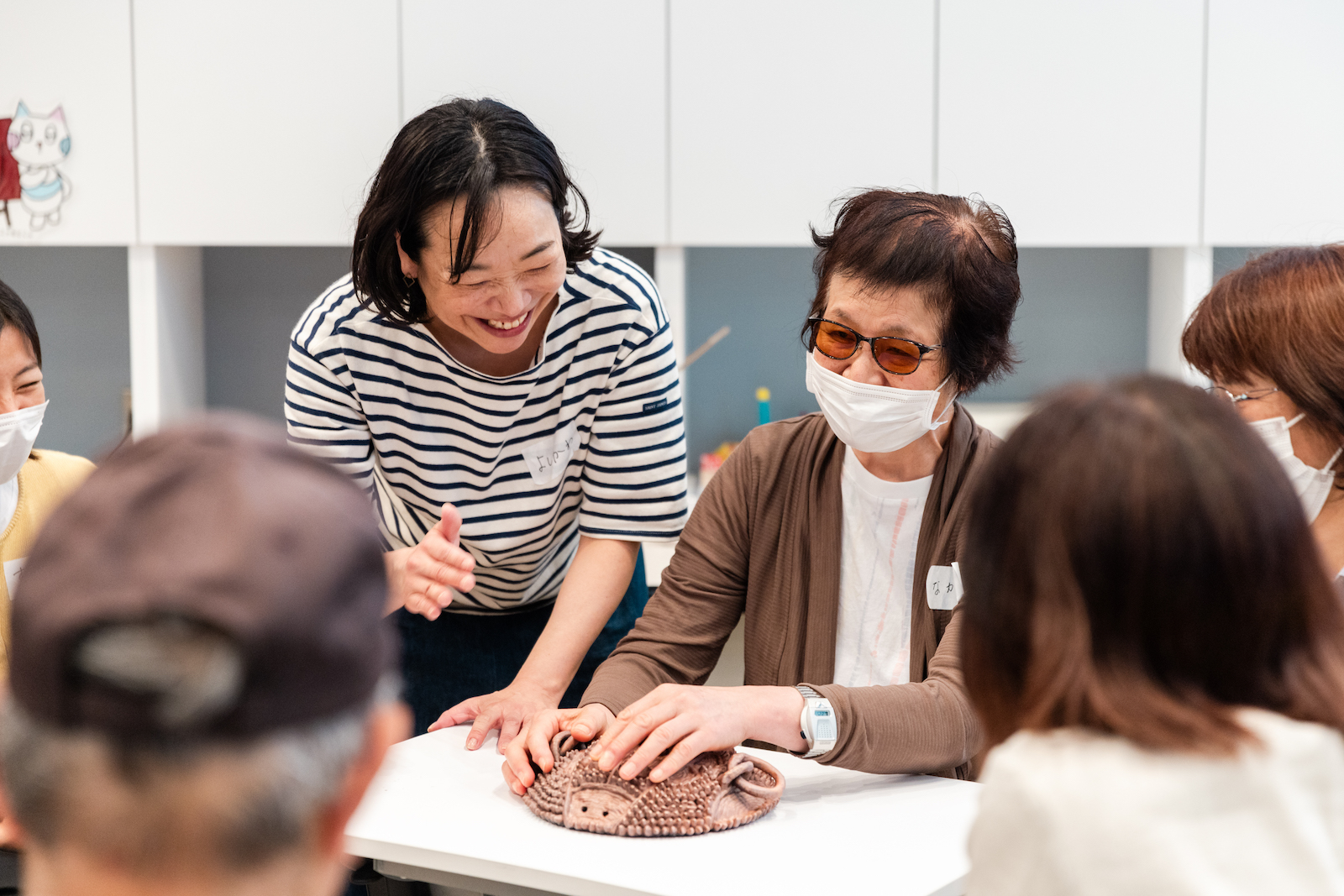 【写真】目の見えにくい参加者のひとりが、机の上に置かれた、無数の突起物に覆われた澤田真一による造形作品を手でさわっている様子。進行役のひとりである滋賀県立美術館学芸員（エデュケーター）の吉川紀子さんをはじめ、参加者がまわりでその様子を笑顔で見守っている。