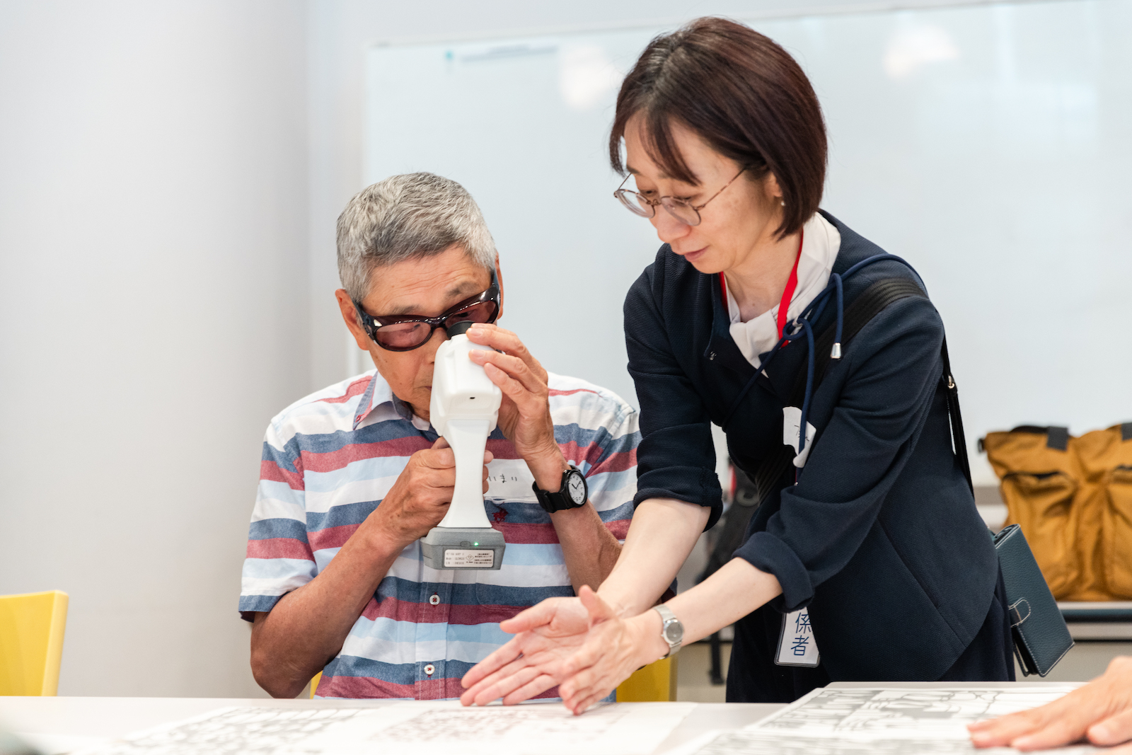 【写真】テーブルのうえに置かれた、幾何学模様のような線を描いた戸來貴規の作品を、「RETISSA ON HAND」で鑑賞する参加者の様子。スタッフが横で鑑賞をサポートしている。